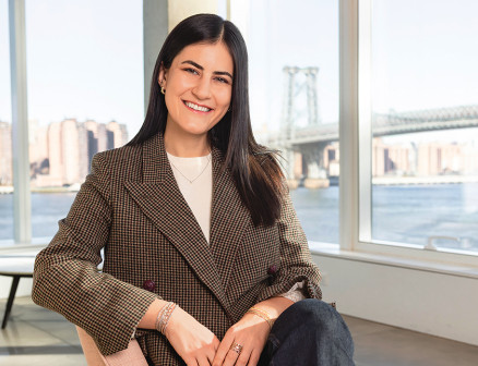 Idil Kara smiles at the camera while wearing a tweed blazer. She sits in a chair in a room with large windows, through which one can see tall buildings along the water and a bridge.