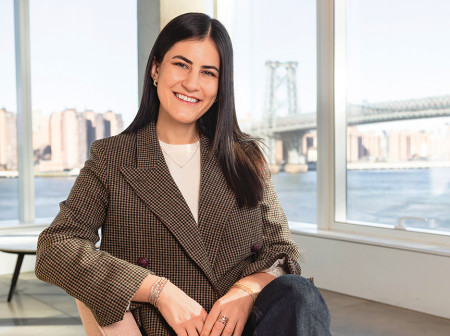 Idil Kara smiles at the camera while wearing a tweed blazer. She sits in a chair in a room with large windows, through which one can see tall buildings along the water and a bridge.