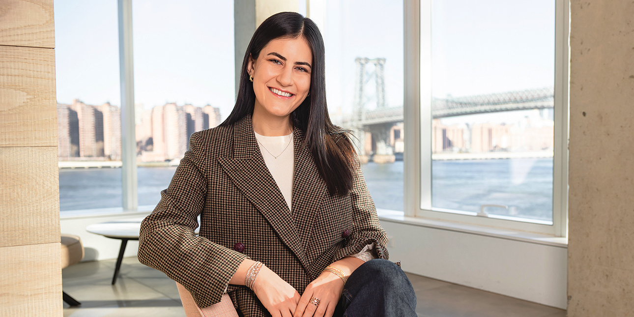 Idil Kara smiles at the camera while wearing a tweed blazer. She sits in a chair in a room with large windows, through which one can see tall buildings along the water and a bridge.