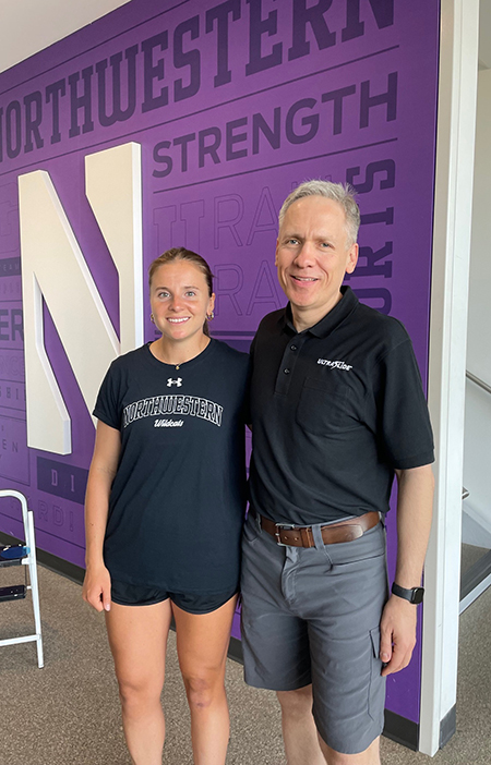 Maddie Zimmer, dressed in athletic clothes, stands to the left of Barry Slotnick, dressed in a black polo and grey shorts. 