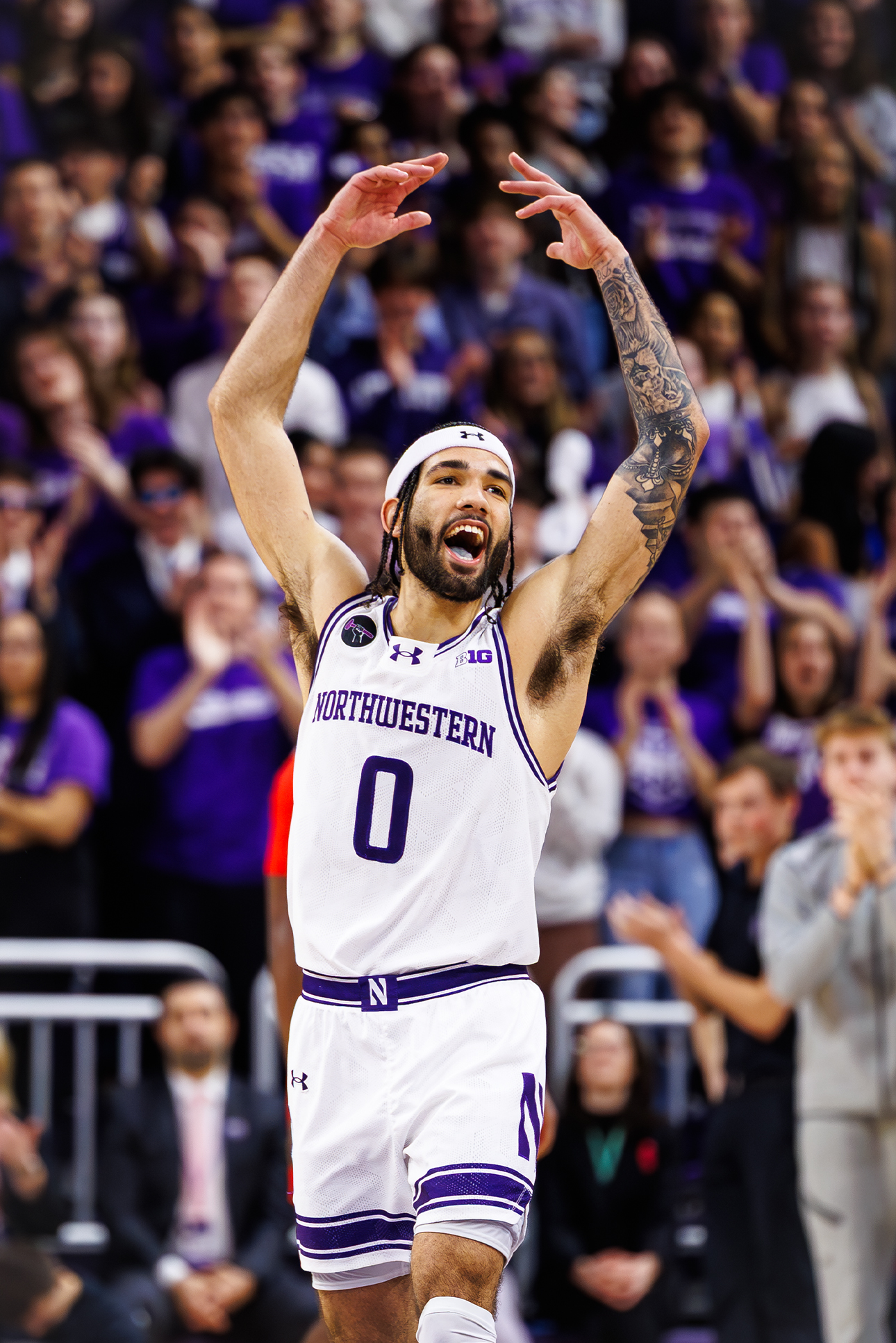 Dressed in a white basketball uniform with purple accents, Boo Buie raises his hands to the air. The student crowd, out of focus, claps in the background.