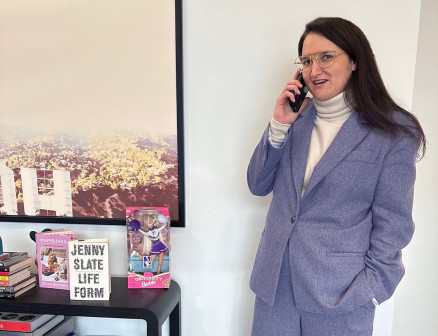 Shauna Perlman stands in a blue suit while talking on the phone next to a table filled with various books and a Northwestern Barbie.