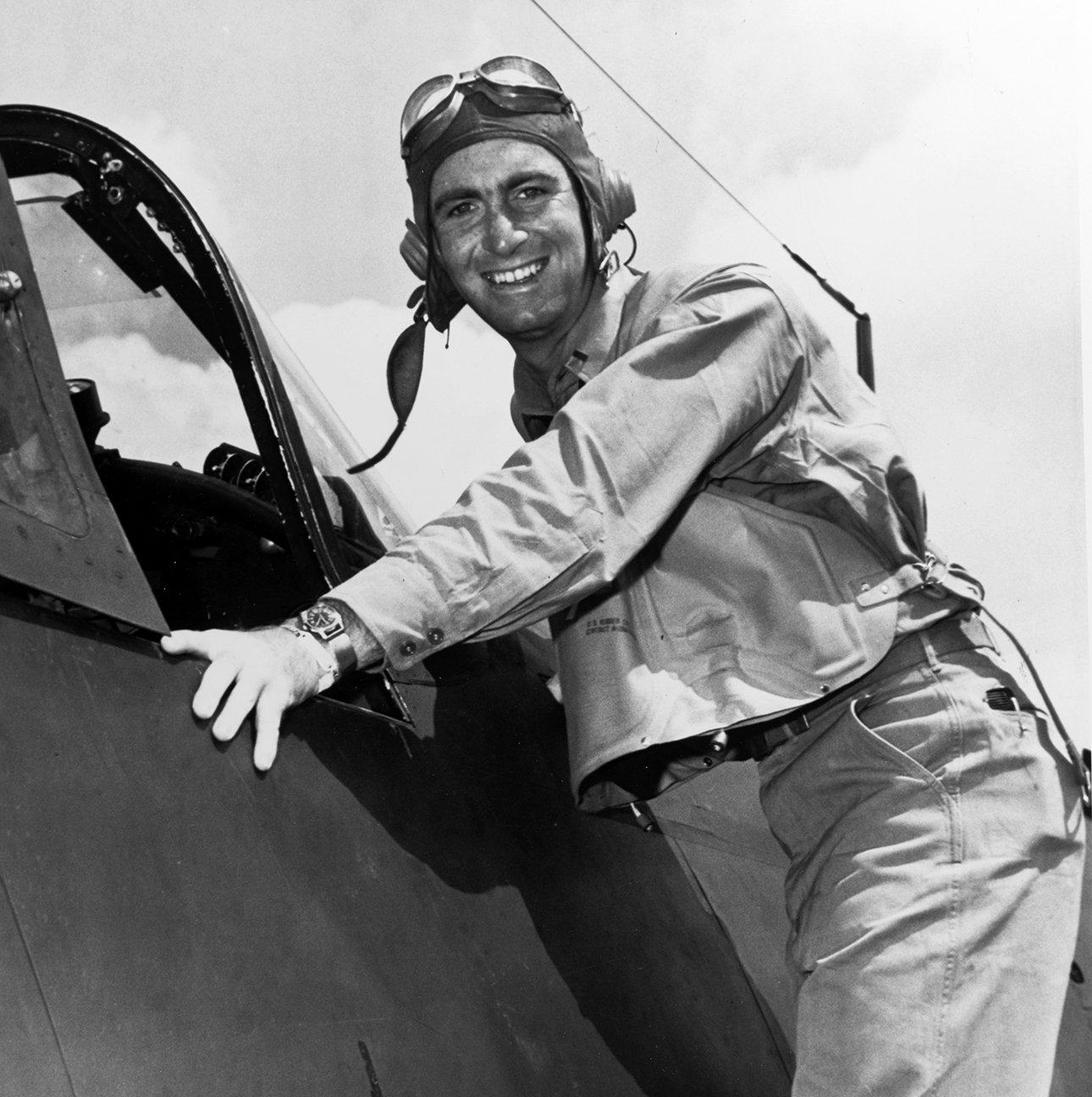 In this black-and-white image, Ike Kepford poses against a VF-17 Corsair plane. He is wearing a light-colored jacket and pants and an aviator’s helmet and goggles on the top of his head. 