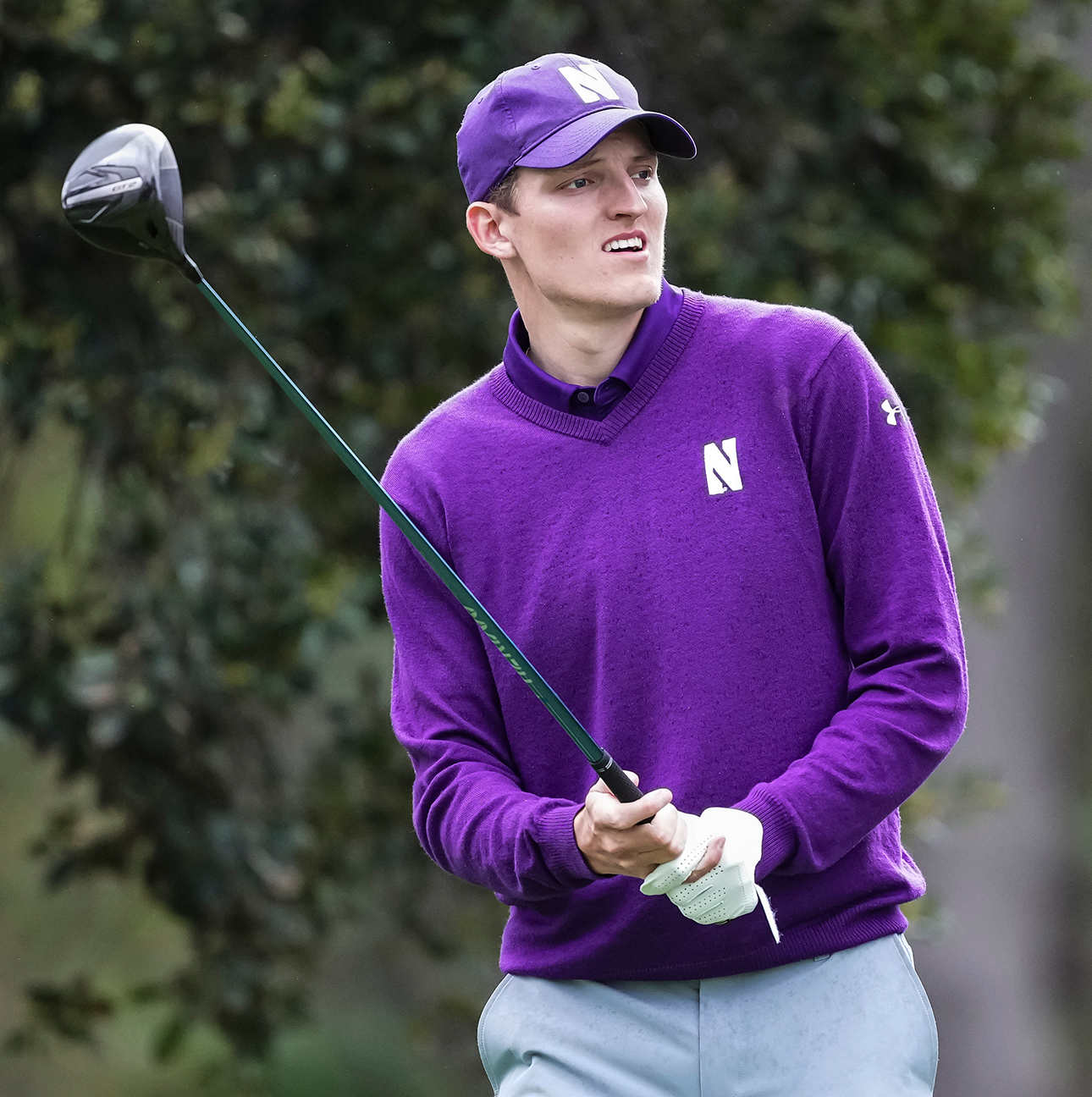 Daniel Svärd, dressed in a purple Northwestern hat, polo and sweater, holds his club up after finishing a swing and watches his shot.