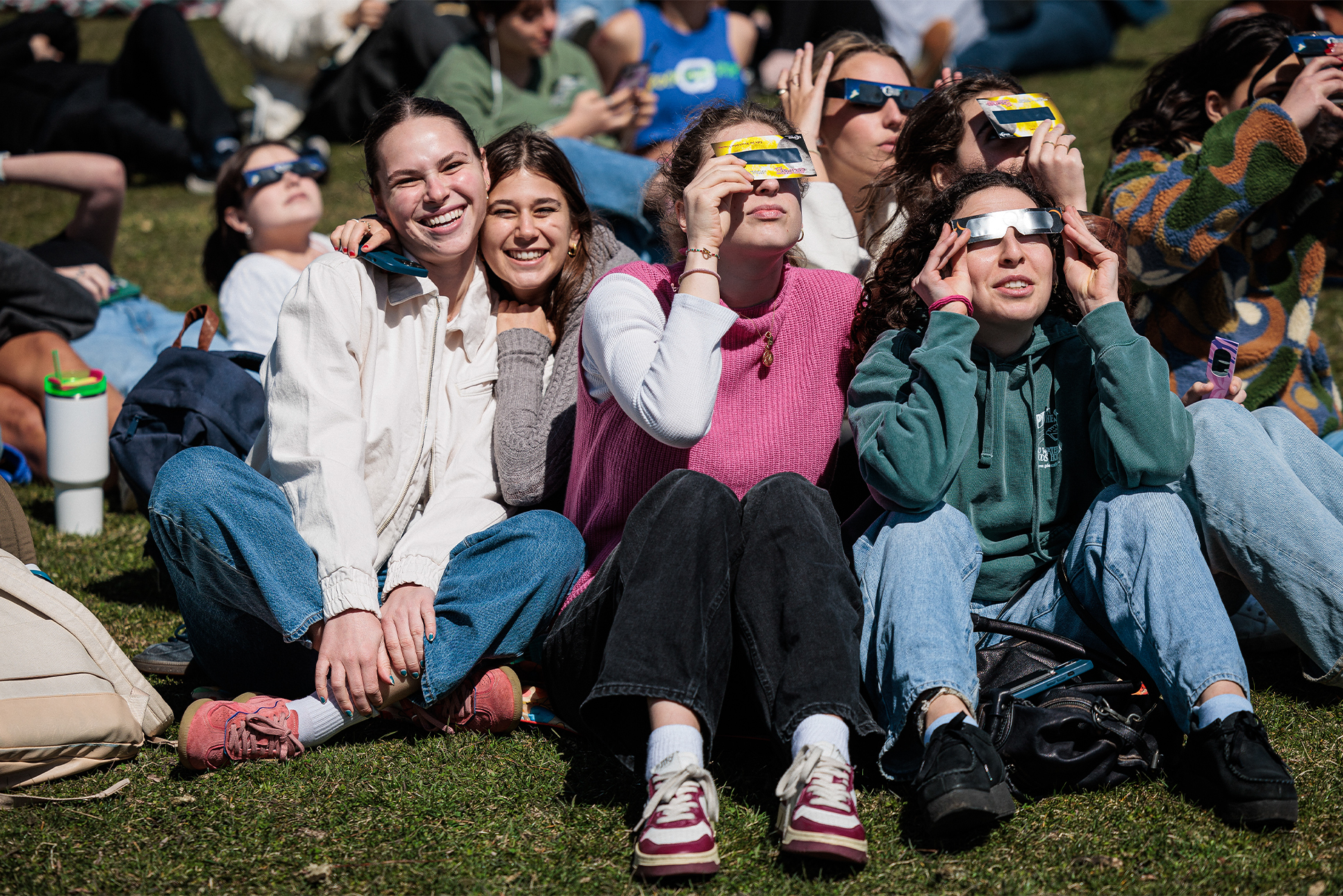 Students sit on the Lakefill, some smiling at the camera and others holding up glasses to view the solar eclipse.