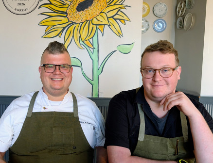 Two men with glasses and green chef aprons are seated next to each other at a restaurant table and smiling for the camera.