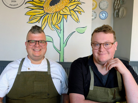 Two men with glasses and green chef aprons are seated next to each other at a restaurant table and smiling for the camera.