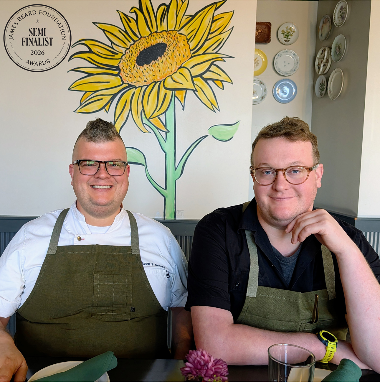Two men with glasses and green chef aprons are seated next to each other at a restaurant table and smiling for the camera.