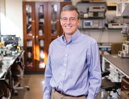 Dressed in a long-sleeved button-down shirt, professor John A. Rogers smiles at the camera with lab equipment in the background.
