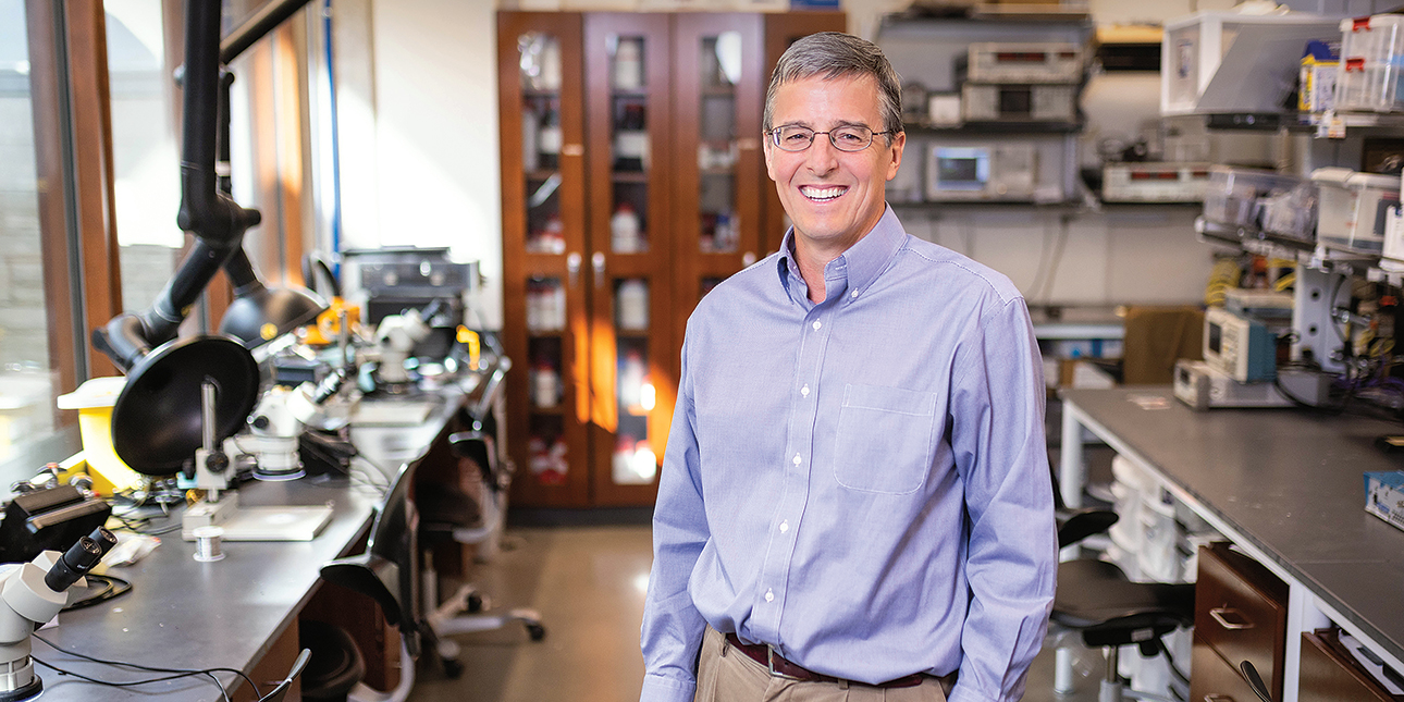 Dressed in a long-sleeved button-down shirt, professor John A. Rogers smiles at the camera with lab equipment in the background.