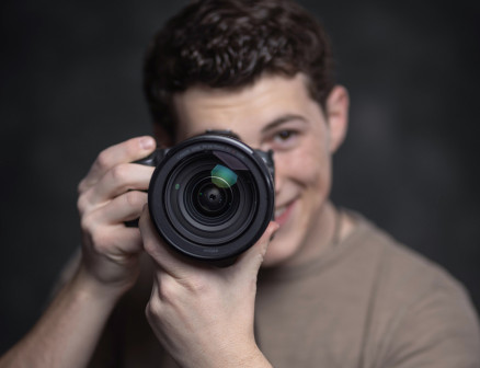 Josh Sukoff, in the background and out of focus, holds up a camera with the lens, in focus, pointed at the reader.