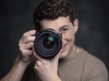 Josh Sukoff, in the background and out of focus, holds up a camera with the lens, in focus, pointed at the reader.