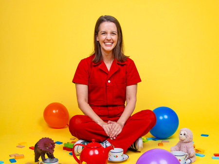 Julie Ritchey sits cross-legged on the ground against a bright yellow background. She is wearing a red jumpsuit and smiling at the camera. Surrounding her are balloons, confetti, teacups and toys.