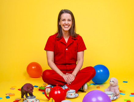 Julie Ritchey sits cross-legged on the ground against a bright yellow background. She is wearing a red jumpsuit and smiling at the camera. Surrounding her are balloons, confetti, teacups and toys.