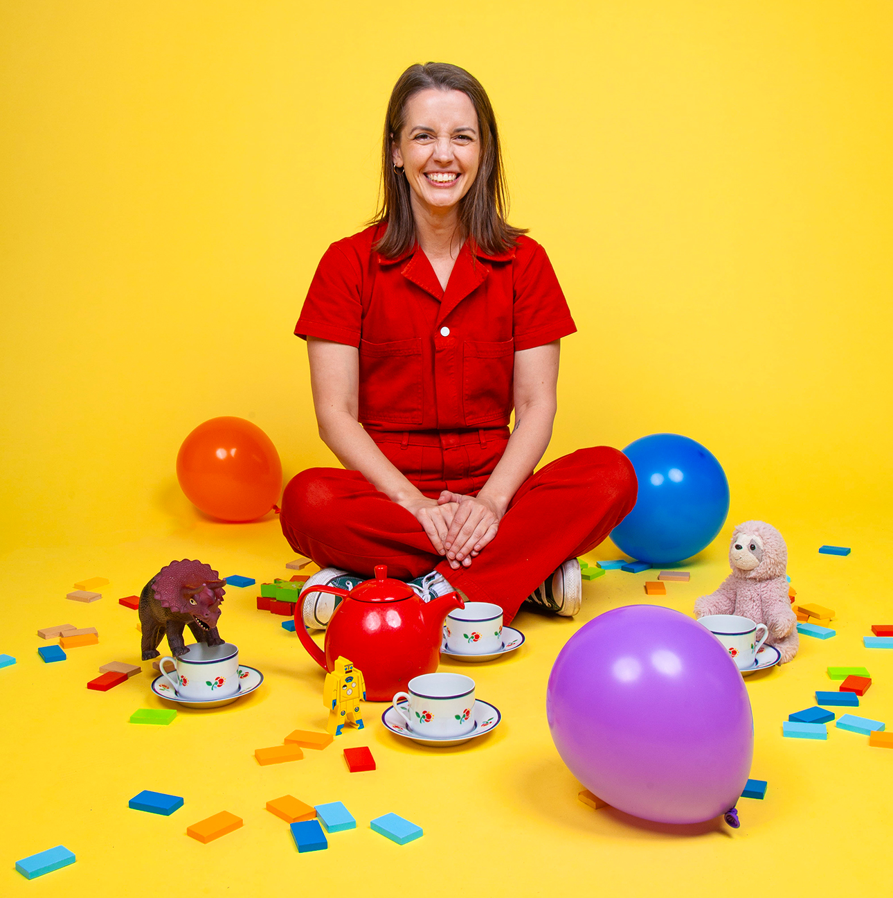 Julie Ritchey sits cross-legged on the ground against a bright yellow background. She is wearing a red jumpsuit and smiling at the camera. Surrounding her are balloons, confetti, teacups and toys.