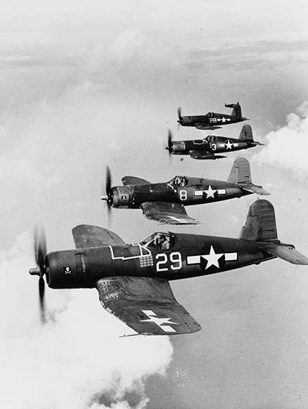 In this black-and-white image, a group of four U.S. Navy Corsair planes, numbered 29, 8, 3 and 28, fly in formation above the clouds and ocean. 
