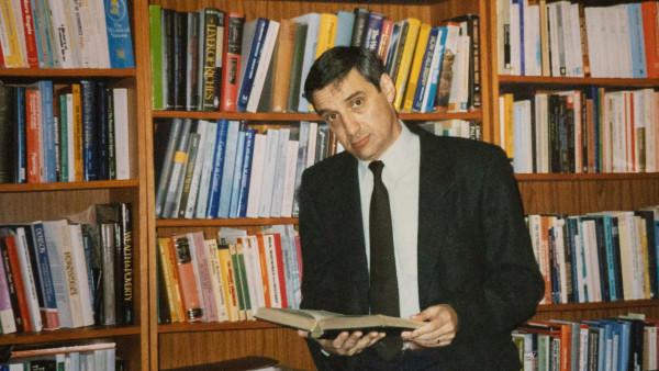 Joel Mokyr stands in front of a bookshelf holding an open book.