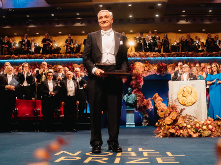 Joel Mokyr, dressed in a tuxedo, stands in an auditorium, smiling and holding his Nobel Prize award. Beneath his feet is a carpet with the words “The Nobel Prize” on it.    Behind Mokyr is a crowd of formally dressed audience members who are giving Mokyr a standing ovation. 