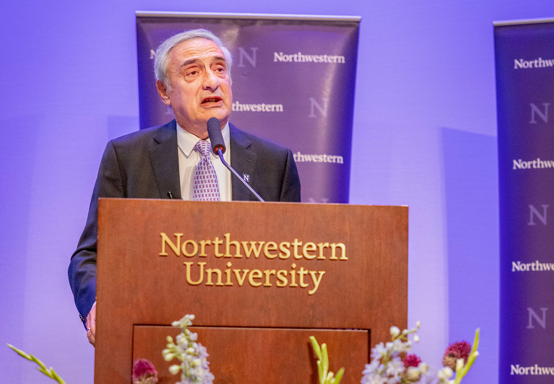 Joel Mokyr is wearing a suit with a purple tie and speaking at a Northwestern University podium. In the foreground are flowers.