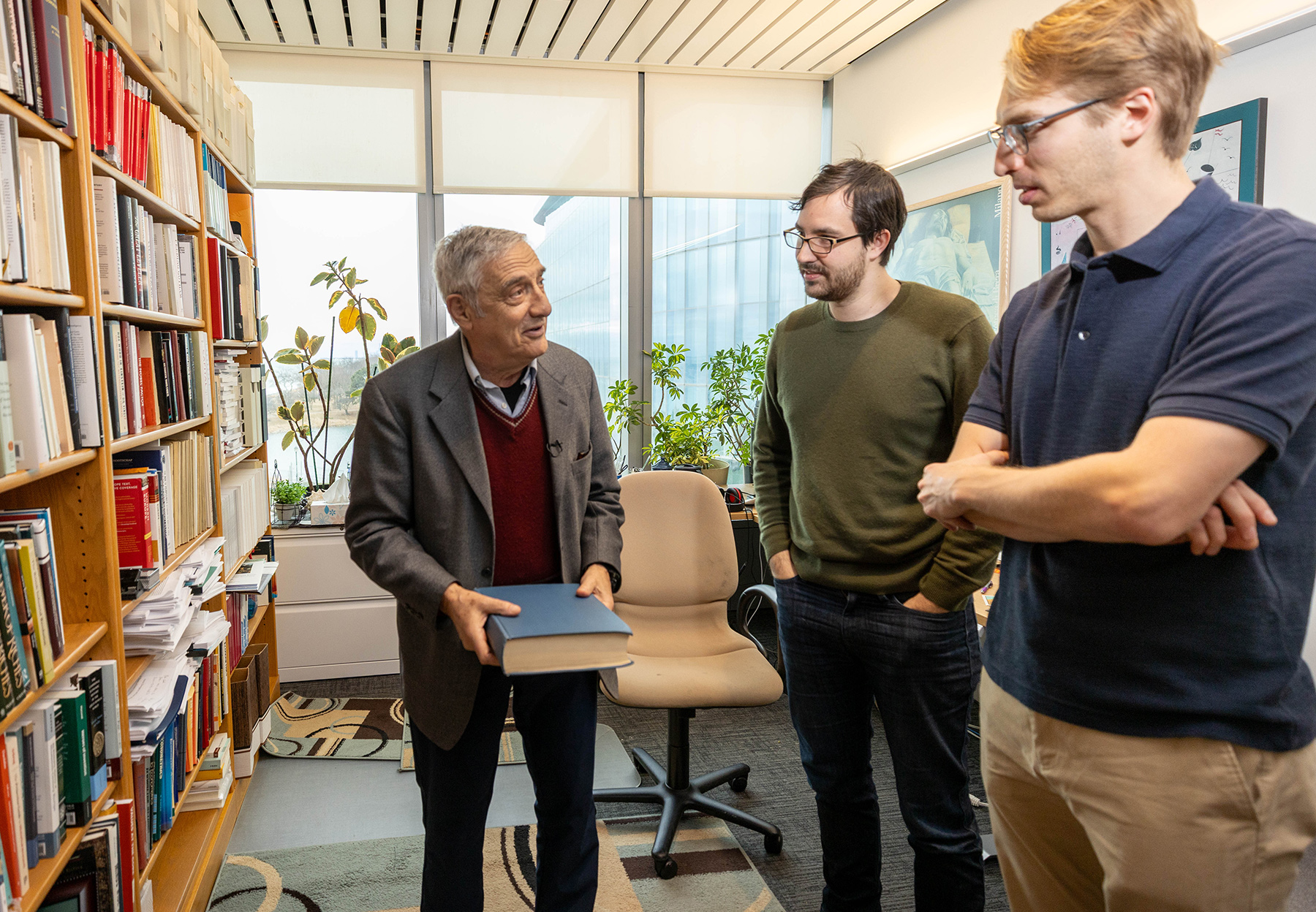 Joel Mokyr stands in his office beside a tall bookshelf that is full of books. He holds a book in his hand while he chats with two students.