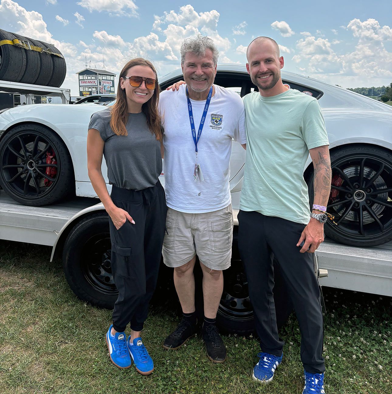 A woman and two men stand outside with their arms around each other in front of a white sports car.