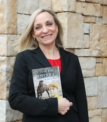 A woman stands against a stone background holding a book tucked under her right arm