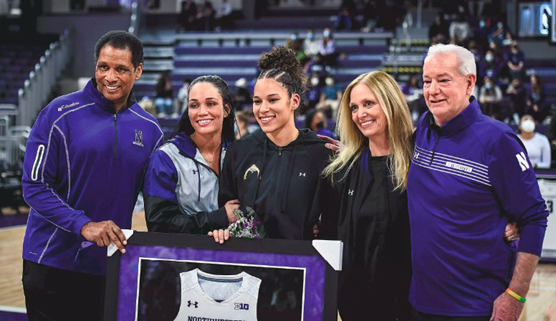Five individuals dressed in Northwestern gear crowd gather together for a photo and hold up Veronica Burton’s framed white Northwestern jersey.