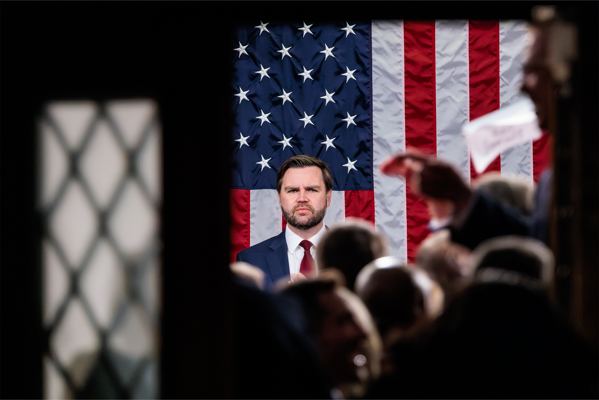 Vice President JD Vance stands facing the camera with an American flag behind him. A crowd, out of focus, stands in front of him.