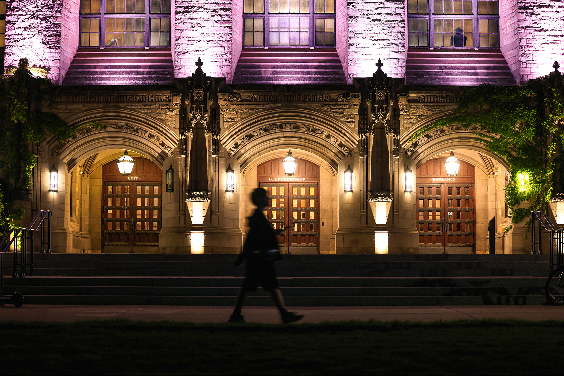 A motion-blurred students walks across the sidewalk in front of a purple-lit Deering Library at night.