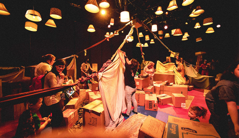 A large room is illuminated with lampshades and lights hanging from the ceiling. Below, children and adults are building forts with cardboard boxes and sheets.