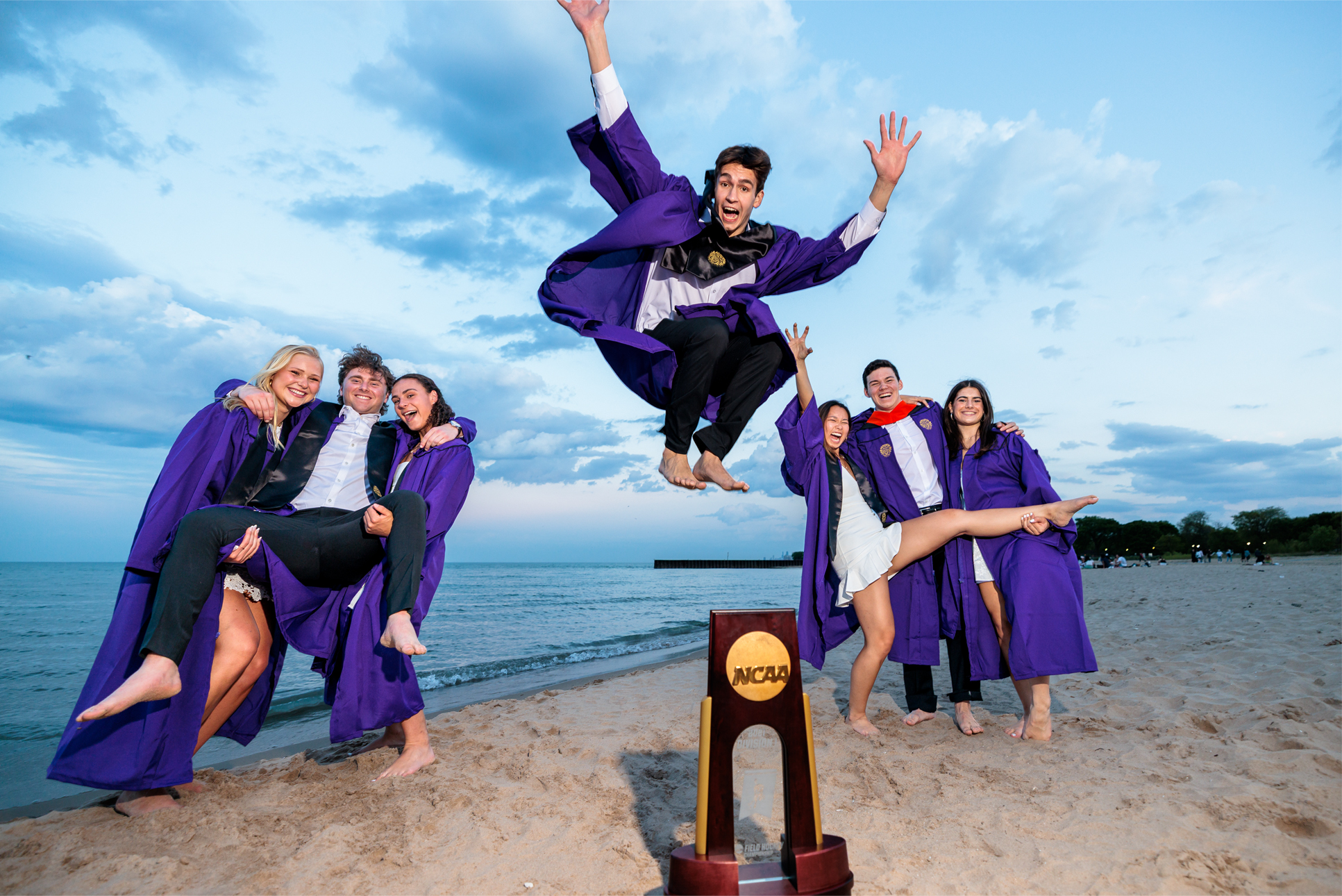 Seven Northwestern seniors pose for a photo on the beach in their purple graduation robes. The one in the middle jumps in the air over an NCAA trophy.