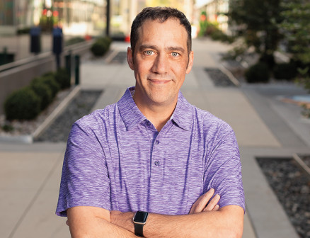 A man in a purple polo shirt smiles on a sidewalk with his arms folded.