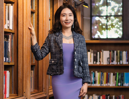 A smiling woman in a purple dress and black and white tweed blazer stands next to a bookshelf in a sunny room.