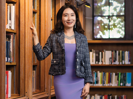 A smiling woman in a purple dress and black and white tweed blazer stands next to a bookshelf in a sunny room.