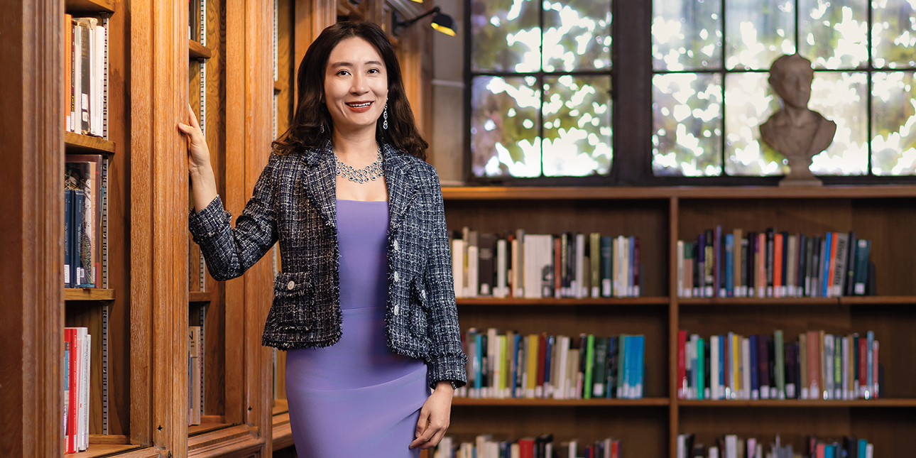 A smiling woman in a purple dress and black and white tweed blazer stands next to a bookshelf in a sunny room.