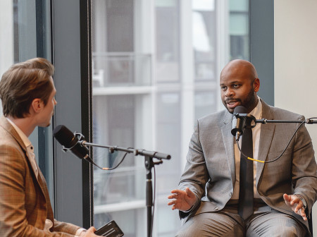 Two men in suits speak to each other while seated at microphones in front of a glass window. 