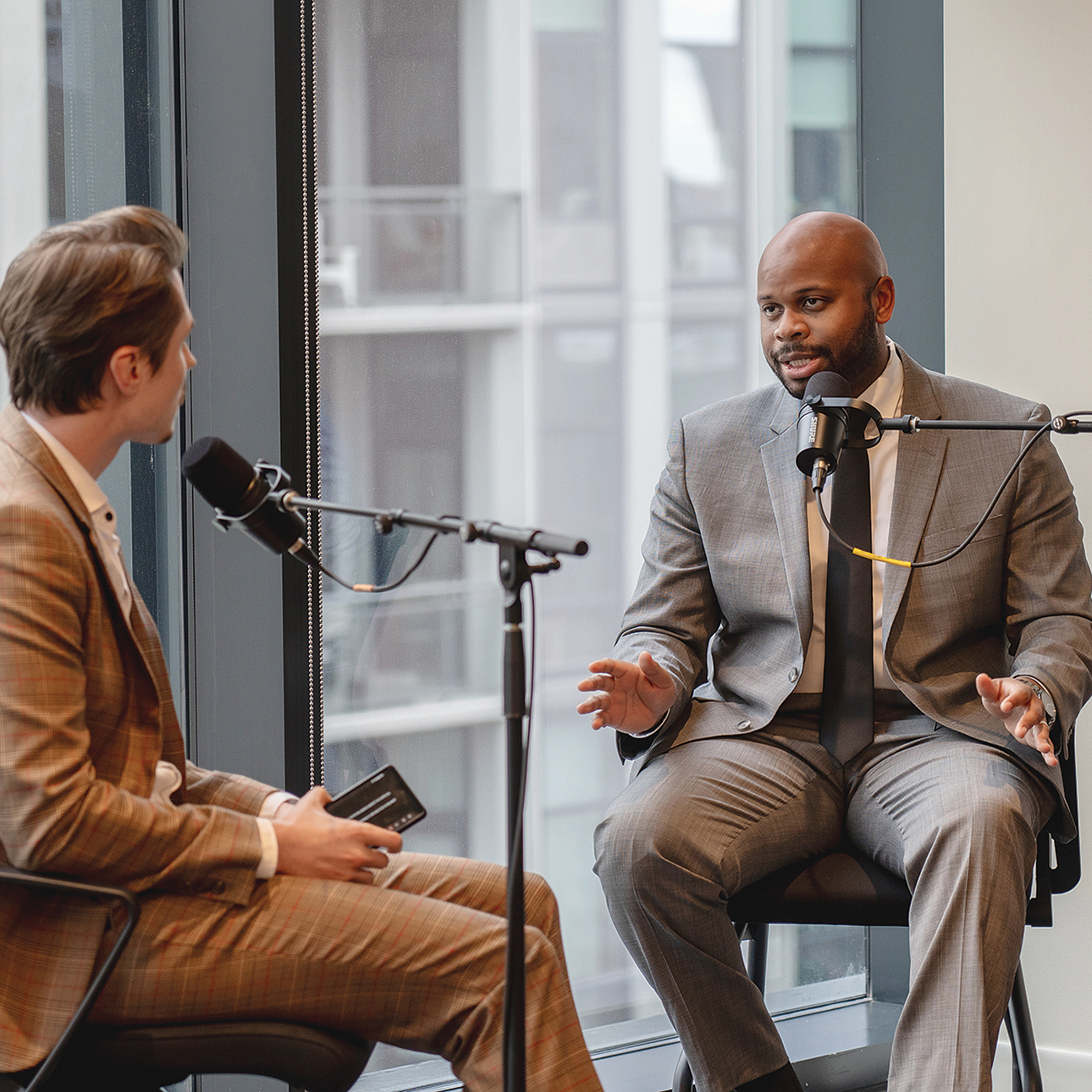 Two men in suits speak to each other while seated at microphones in front of a glass window. 
