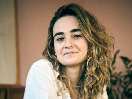 A woman with curly blonde hair wearing a white button-down shirt and blue jeans leans on a counter for her arms crossed.