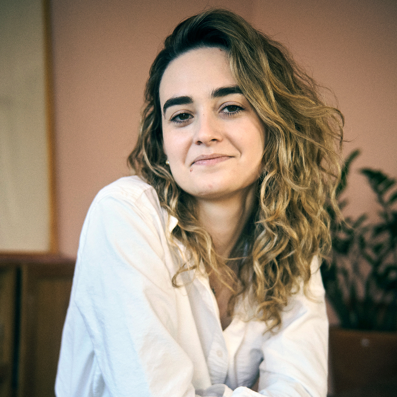 A woman with curly blonde hair wearing a white button-down shirt and blue jeans leans on a counter for her arms crossed.
