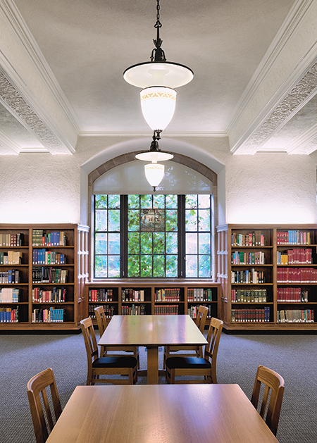 A room with walls of bookshelves and wooden tables and chairs at the center and a window in the background.