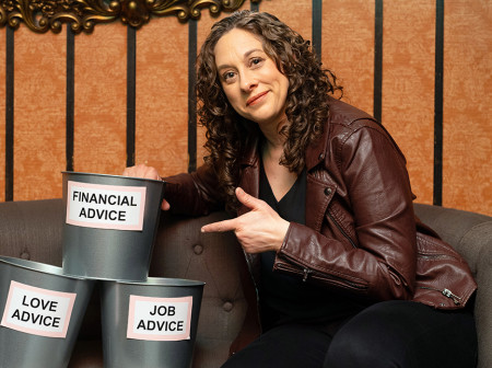 Jenny Hagel wears a brown leather jacket and black pants. She is sitting on a gray couch, pointing to a stack of three metal buckets beside her that are labeled “Financial Advice,” “Love Advice” and “Job Advice.”