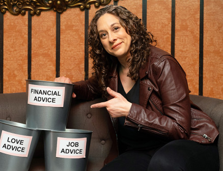 Jenny Hagel wears a brown leather jacket and black pants. She is sitting on a gray couch, pointing to a stack of three metal buckets beside her that are labeled “Financial Advice,” “Love Advice” and “Job Advice.”