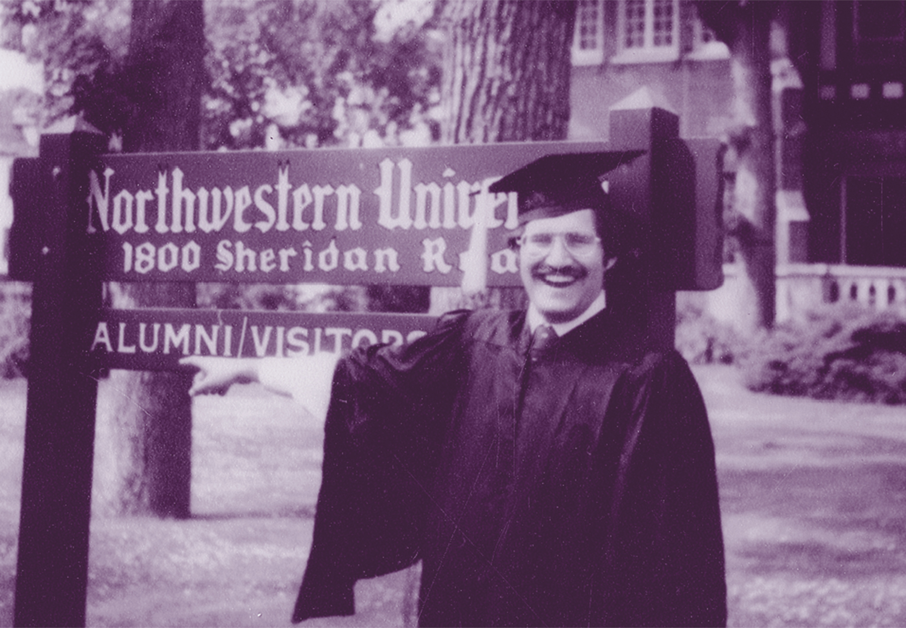 A black-and-white photo of a young Richard Kind wearing his graduation robe and mortarboard and grinning in front of a sign that says Northwestern University.