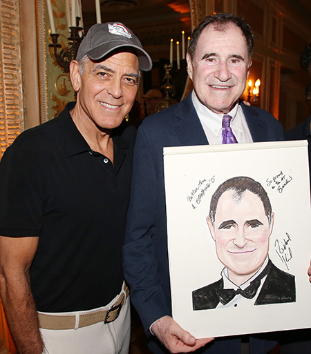 George Clooney and Richard Kind stand beside each other and smile at the camera while Kind holds a caricature drawing of himself.