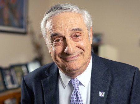 Joel Mokyr, wearing a purple Northwestern tie and dark gray suit, smiles at the camera in front of a blurred background.