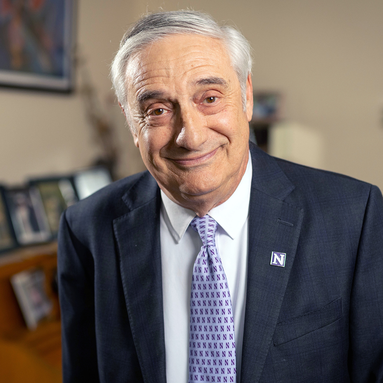 Joel Mokyr, wearing a purple Northwestern tie and dark gray suit, smiles at the camera in front of a blurred background.