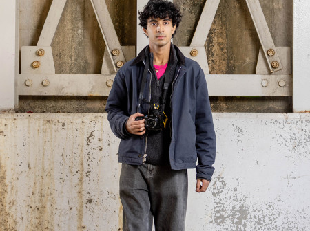 Nikash Khanna stands in front of a concrete underpass. He wears dark jeans and a dark jacket and holds his camera in one hand.