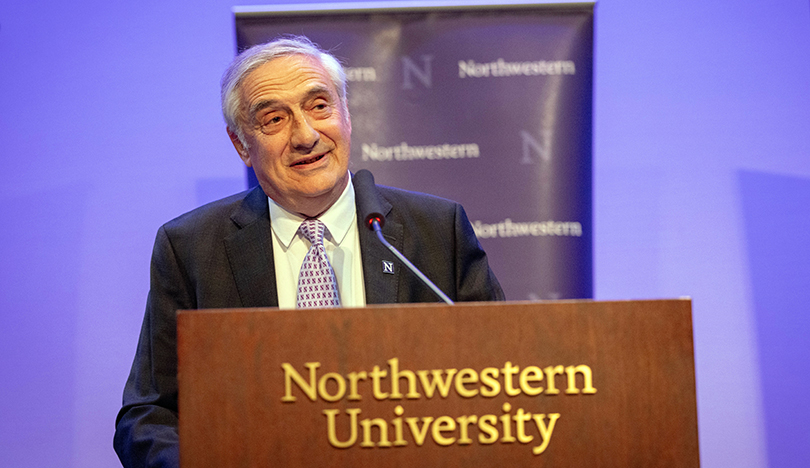 Joel Mokyr stands behind a brown podium with “Northwestern University” on it and delivers a speech; a purple Northwestern banner hangs behind him.