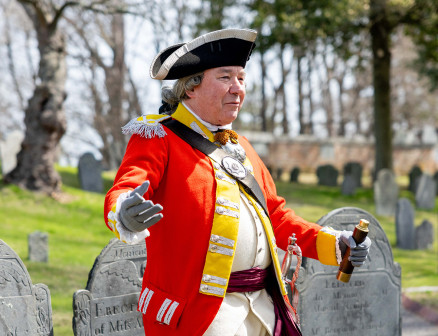 A man reenacting the American Revolution wears a red coat and black hat and is moving his arms in front of tombstones at a cemetery. 