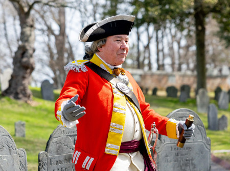 A man reenacting the American Revolution wears a red coat and black hat and is moving his arms in front of tombstones at a cemetery. 